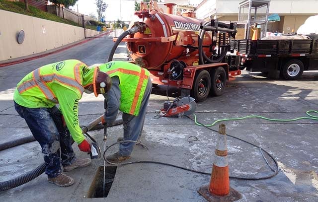 Two workers in yellow safety vests performing sewer maintenance with vacuum truck in background